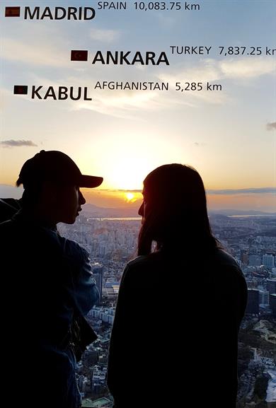 The picture shows a woman and a man, apparently a young couple, watching the sunset from the Namsan Tower in Seoul.